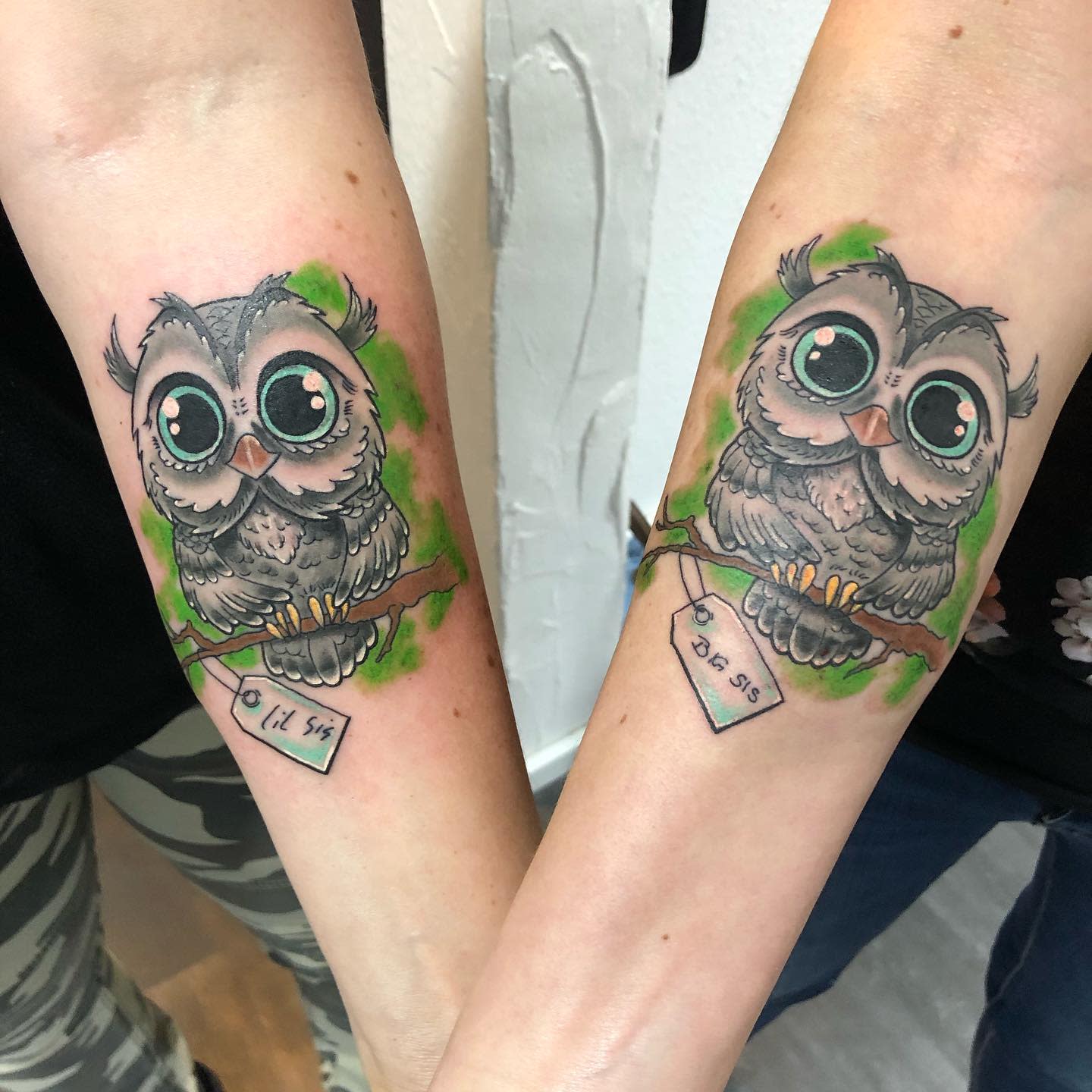 Matching owl tattoos on forearm perched on branches with green background and date tags